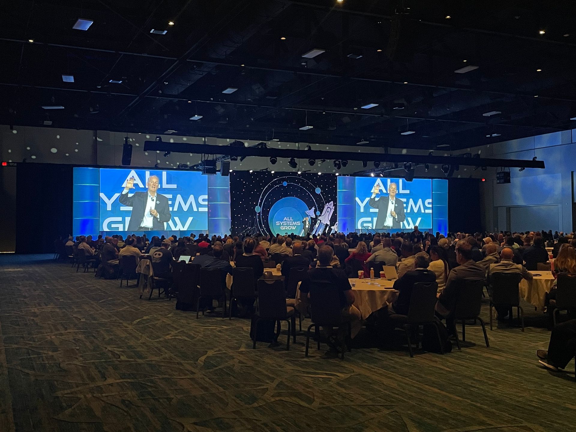 A speaker presents to a large audience in a dimly lit convention hall with large screens displaying 