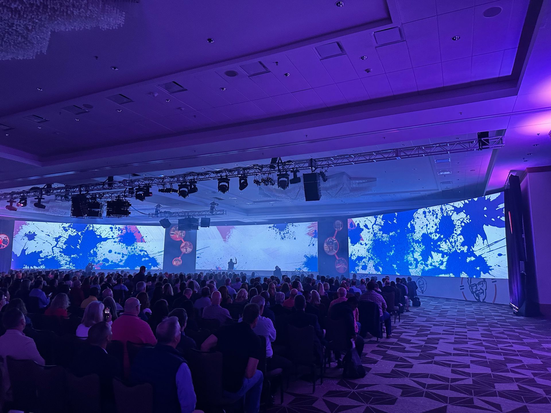 A large audience sits in a dimly lit conference hall, facing a wide, bright screen with blue abstract graphics.