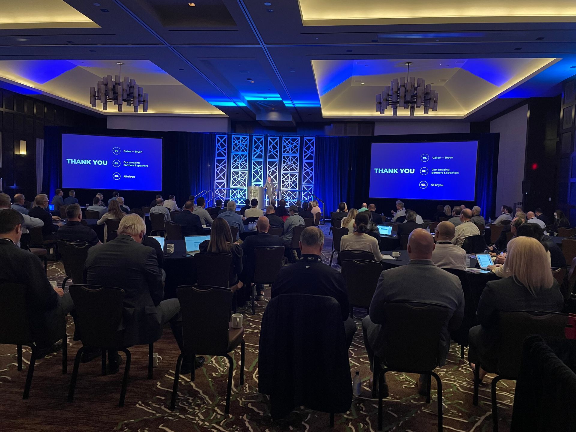 A conference room with a stage, two large screens, and an audience seated at tables facing the presentation.