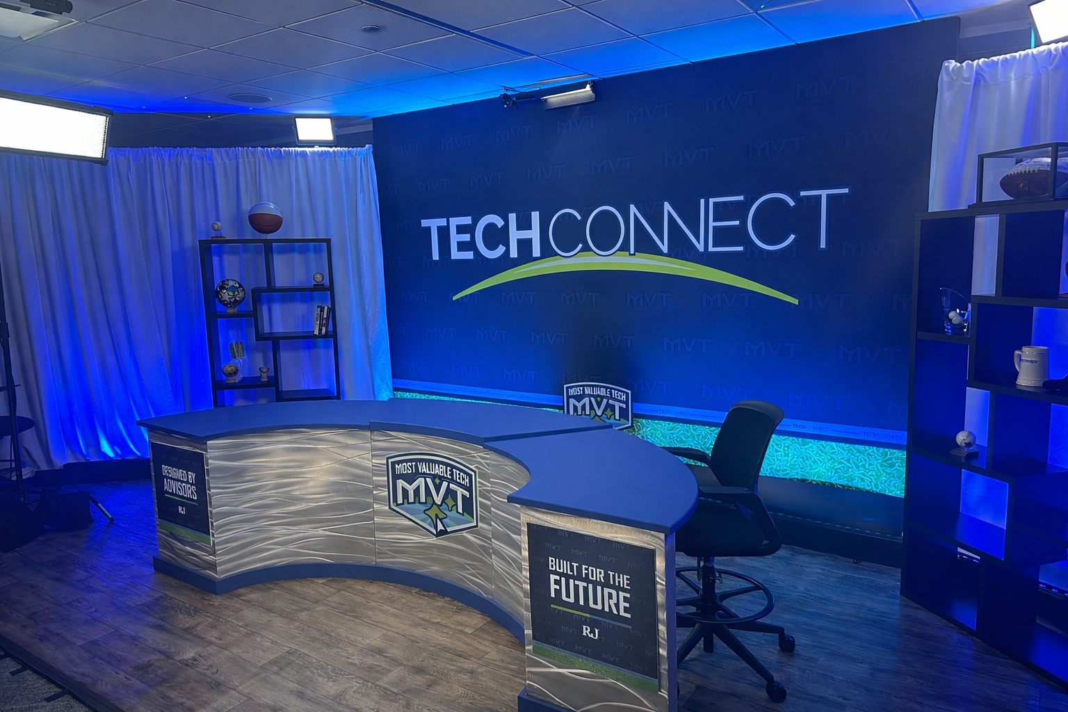 A TV studio set featuring a curved desk, a desk chair, and a blue wall with the 