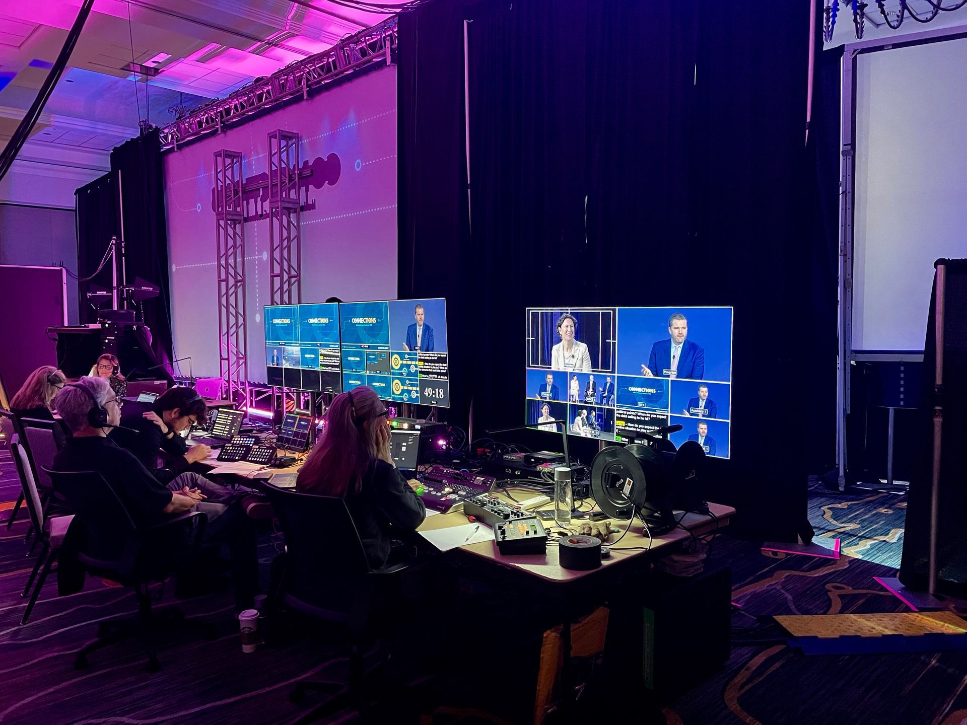 A technical crew sits at a production desk in a darkened room, viewing multiple broadcast monitors for a live event.