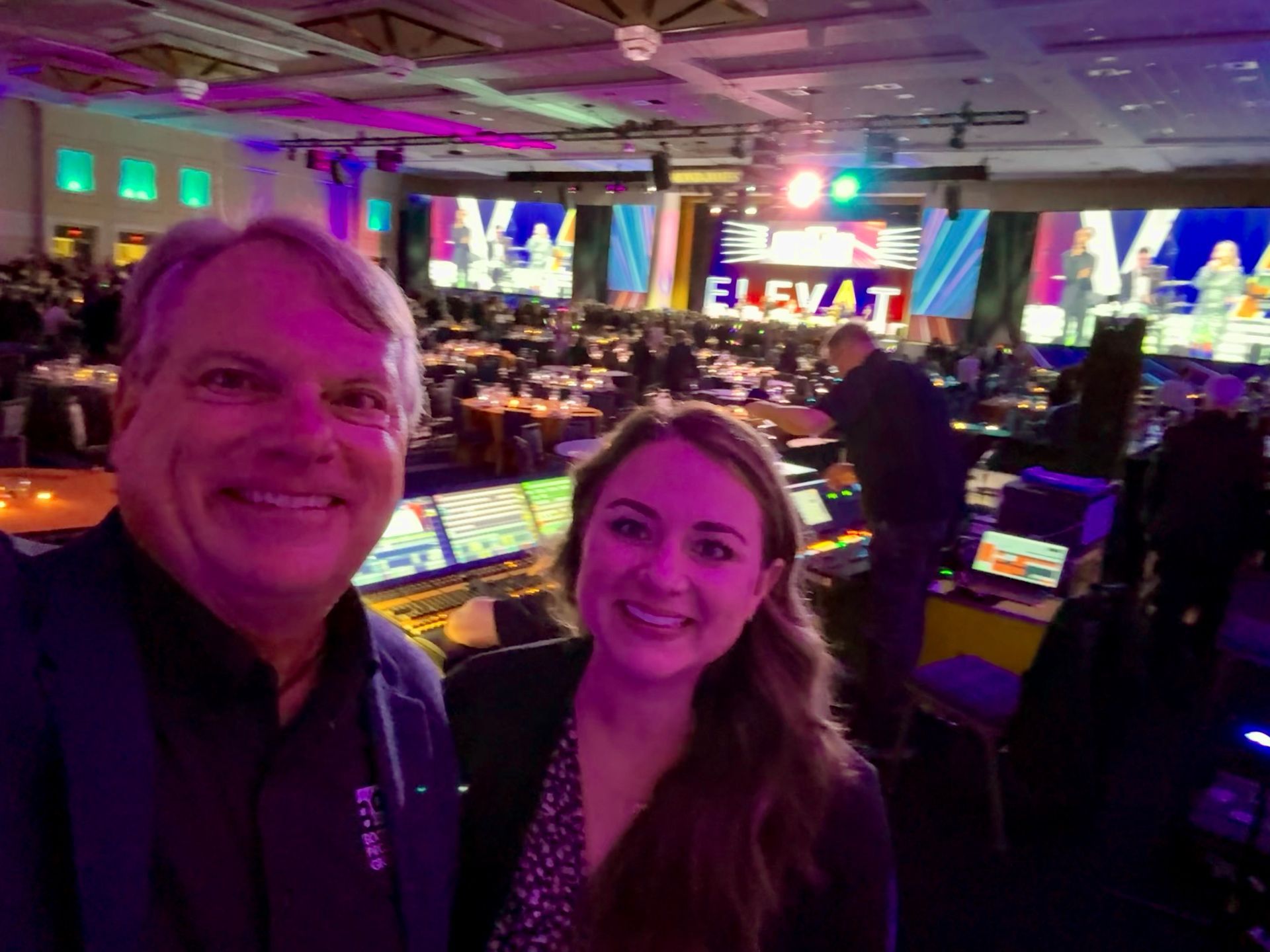 A man and woman pose for a selfie in a large, dimly lit conference hall with stage lighting and event screens.