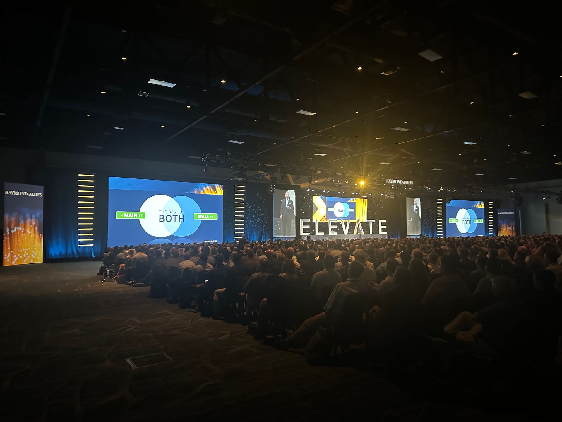 A large audience sits in a dark, expansive convention hall facing a stage with large screens displaying