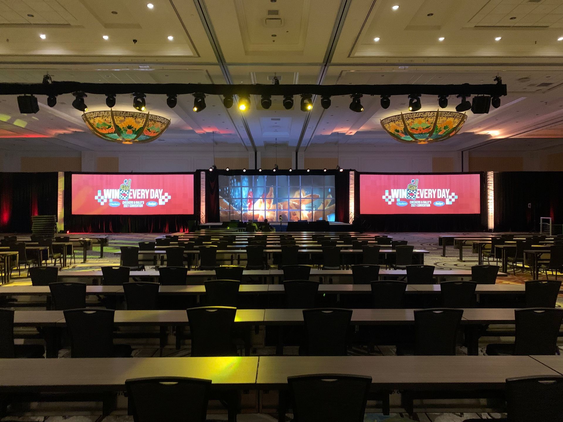 A large conference room with rows of tables facing a stage featuring three screens displaying event branding.