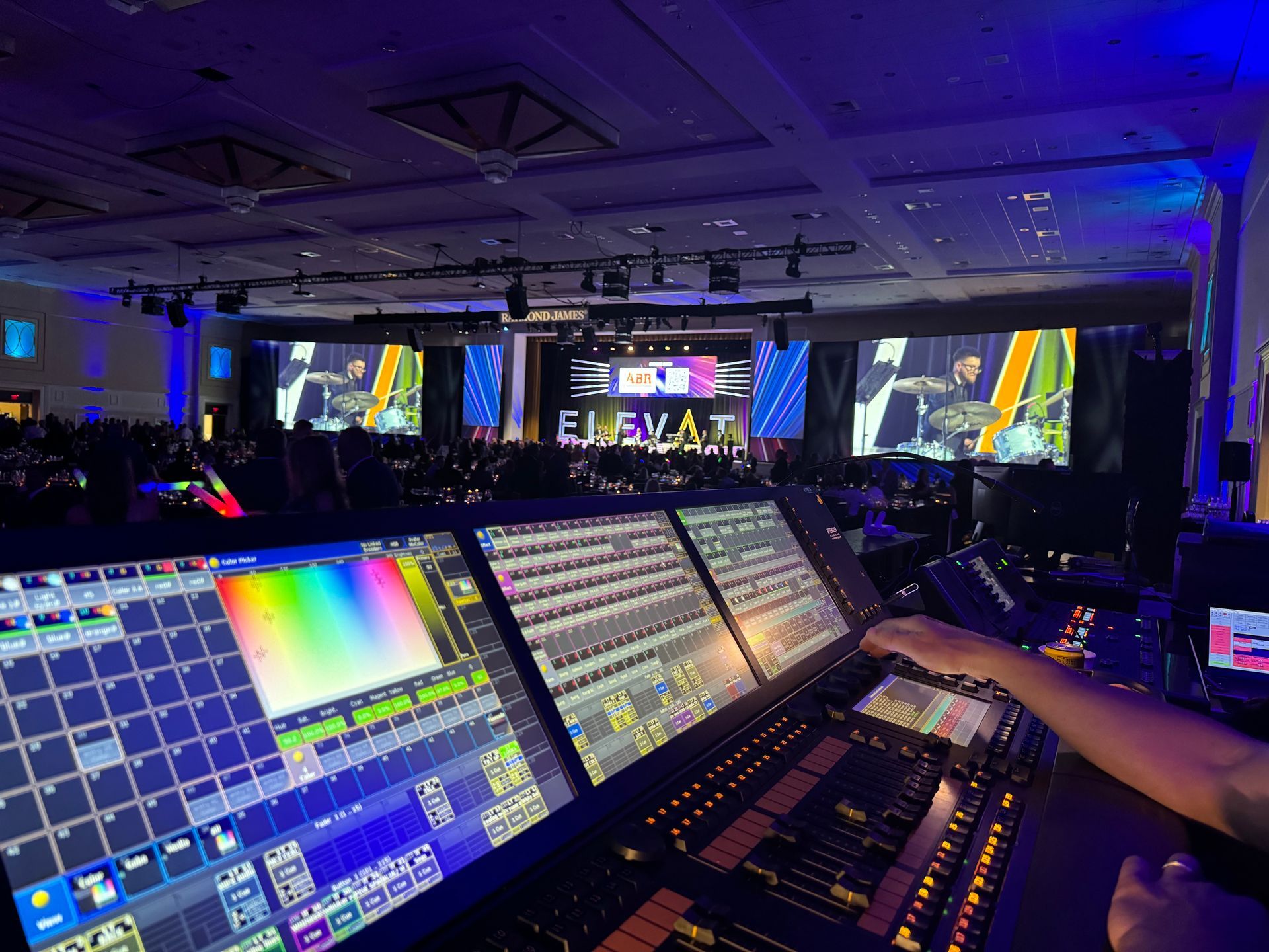 A lighting control console showing colorful displays, positioned in front of a stage at a dimly lit conference event.