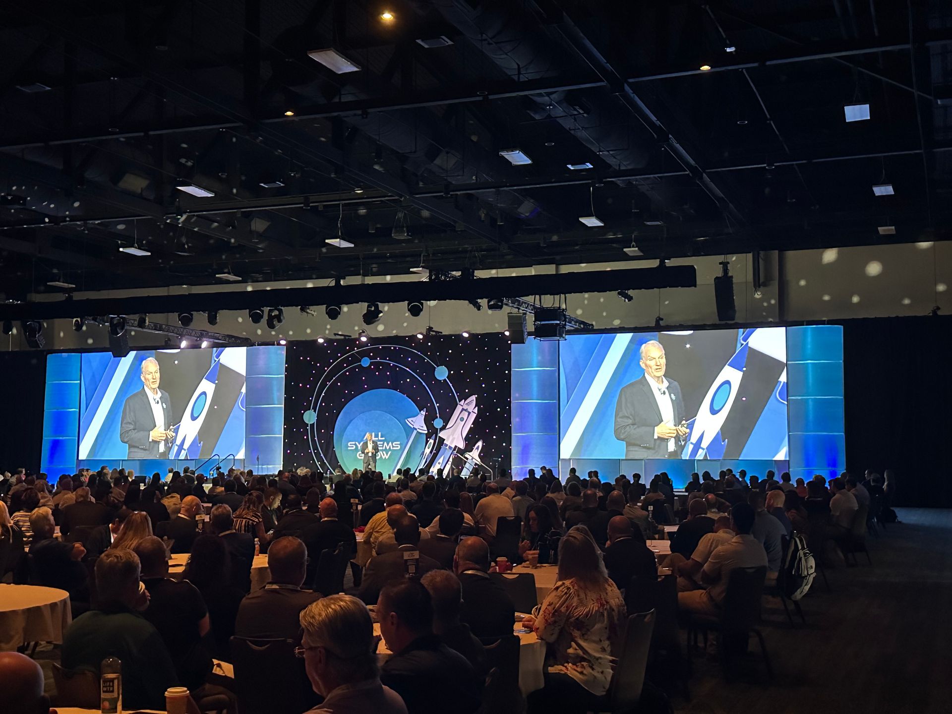 A speaker presents on a large stage in a dimly lit ballroom before a seated audience at round tables.