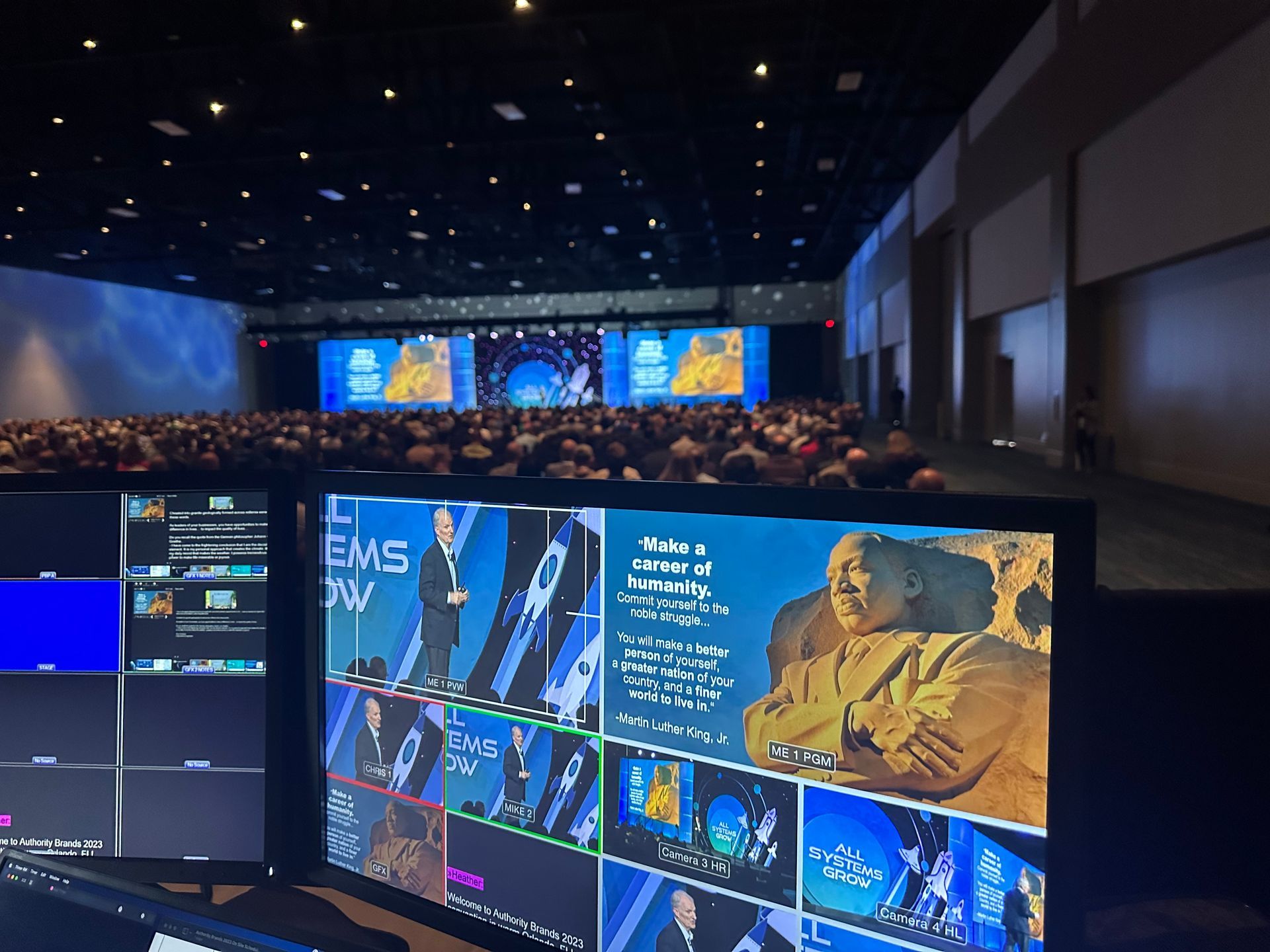 A video control booth overlooking a large, crowded auditorium with multiple screens displaying event speakers and graphics.