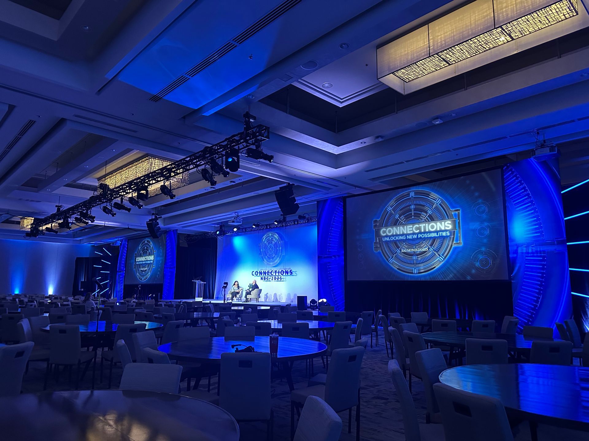An expansive hotel ballroom arranged for a conference with blue lighting, round tables, and large digital screens.