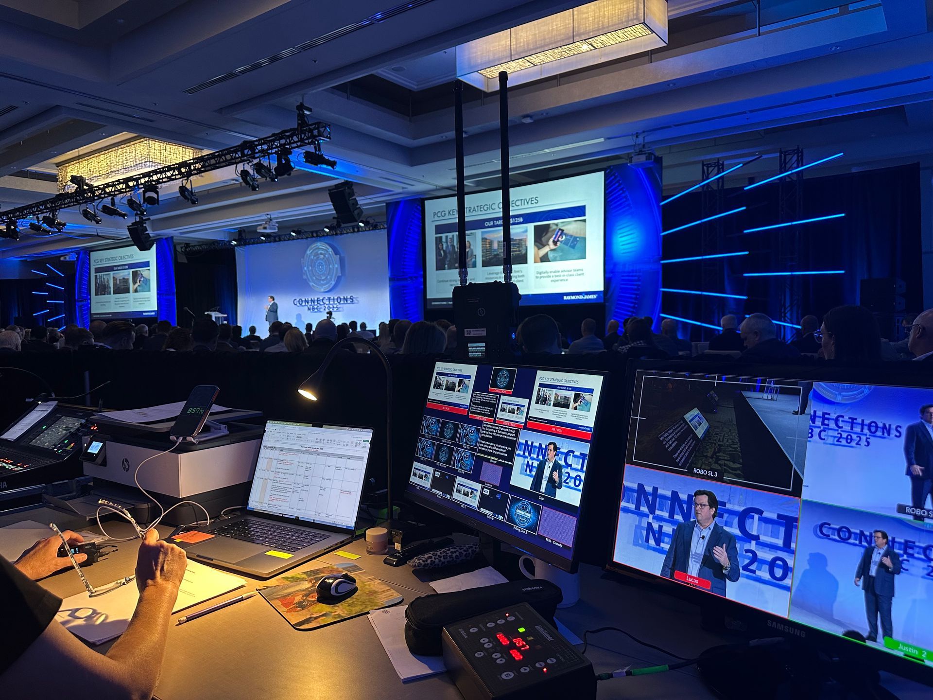 Technicians at a control desk monitor screens displaying a live presentation at an indoor conference venue.