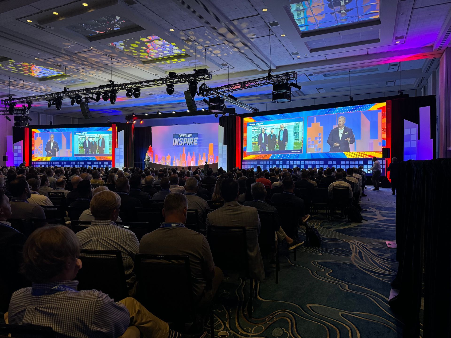 A large audience sits in a dimly lit conference hall watching a speaker on two giant screens.