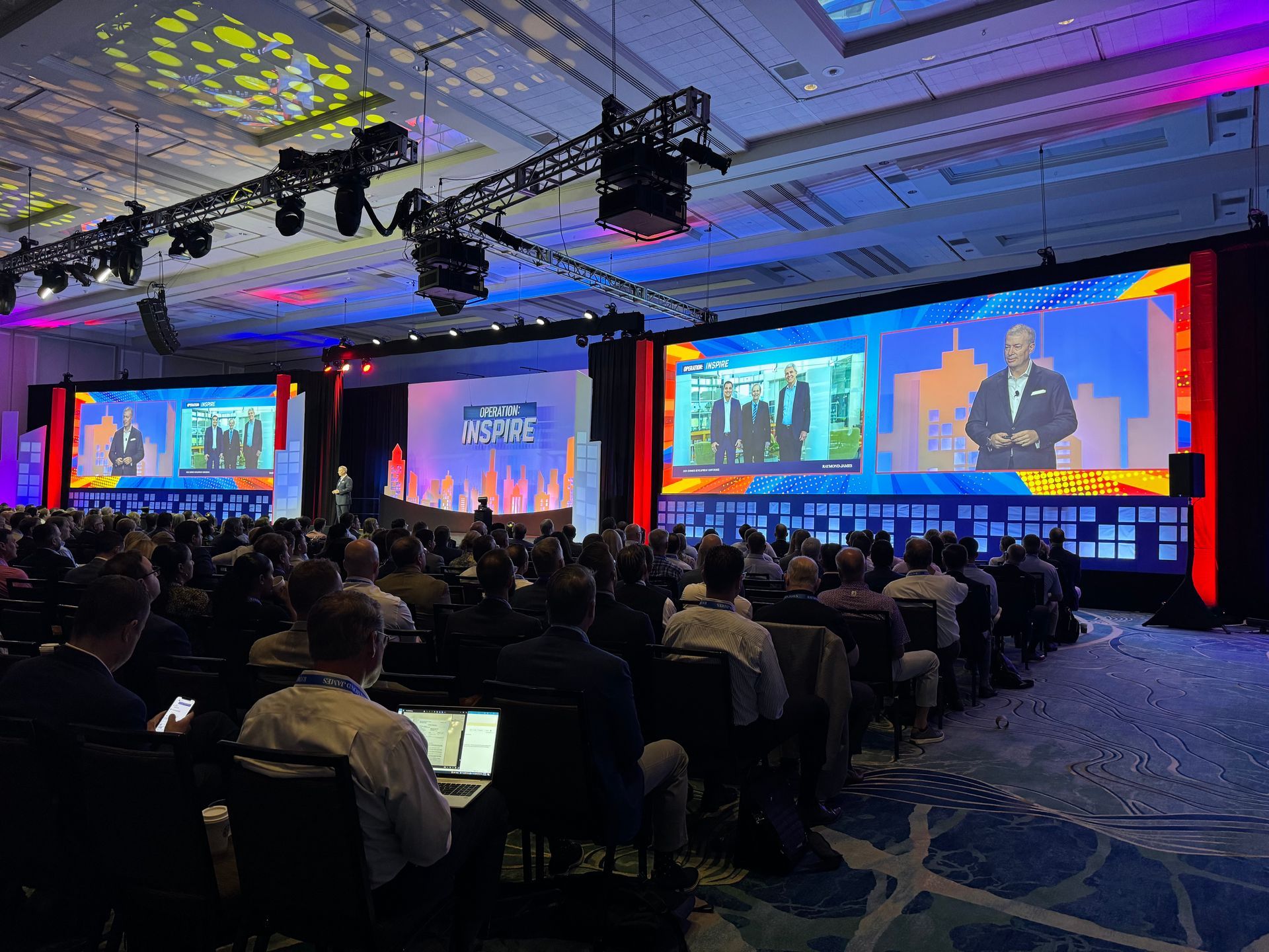 A large audience watches a speaker on stage at a brightly lit indoor conference with multiple projection screens.