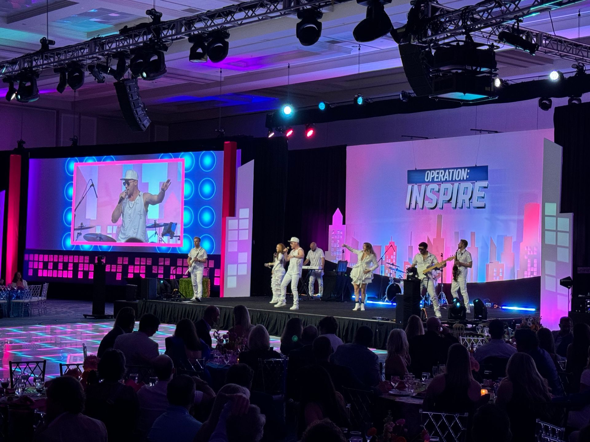 A band in matching white outfits performs on a stage under bright lights, with a large 