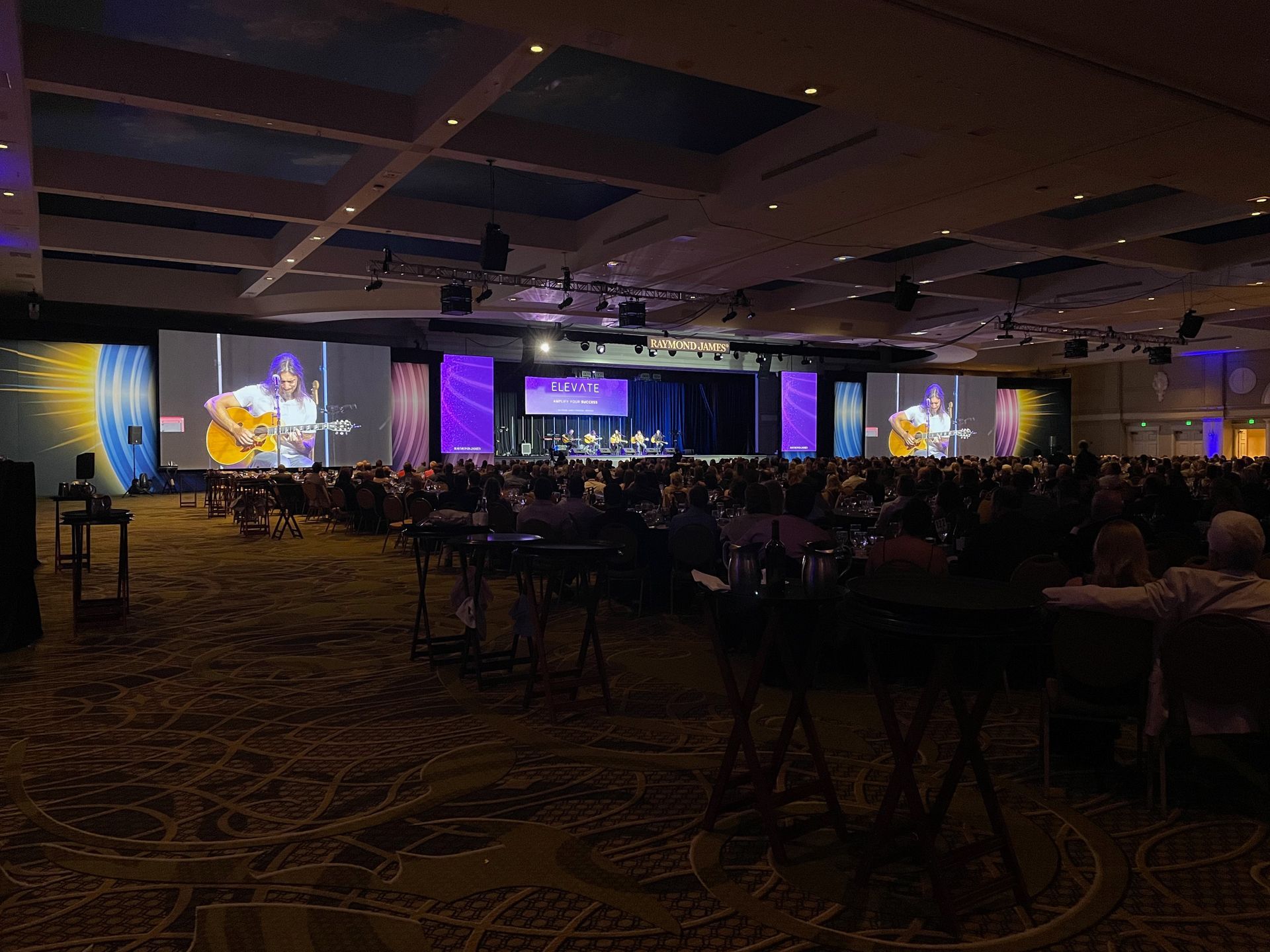 A stage with musicians performing for a large, seated audience in a dimly lit, carpeted conference ballroom.