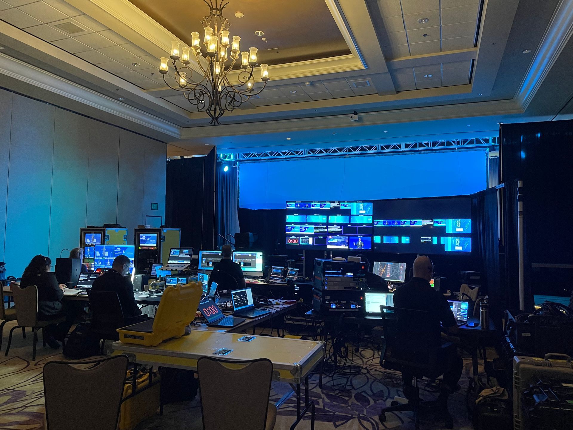 A production crew works at a control desk in a large hotel conference room with a blue-lit stage and large video screens.
