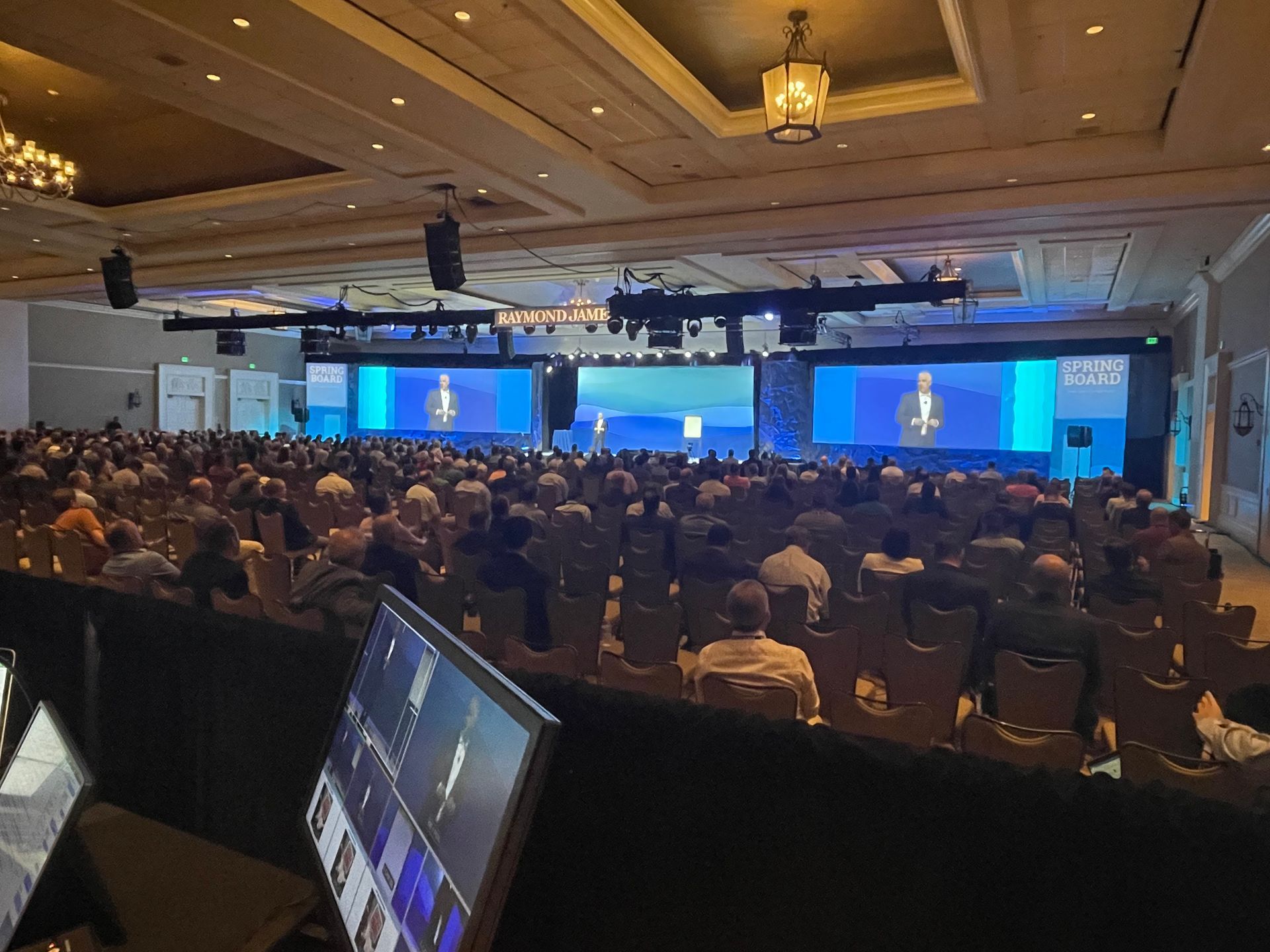 A large conference room filled with seated attendees facing three screens displaying a speaker on a stage.