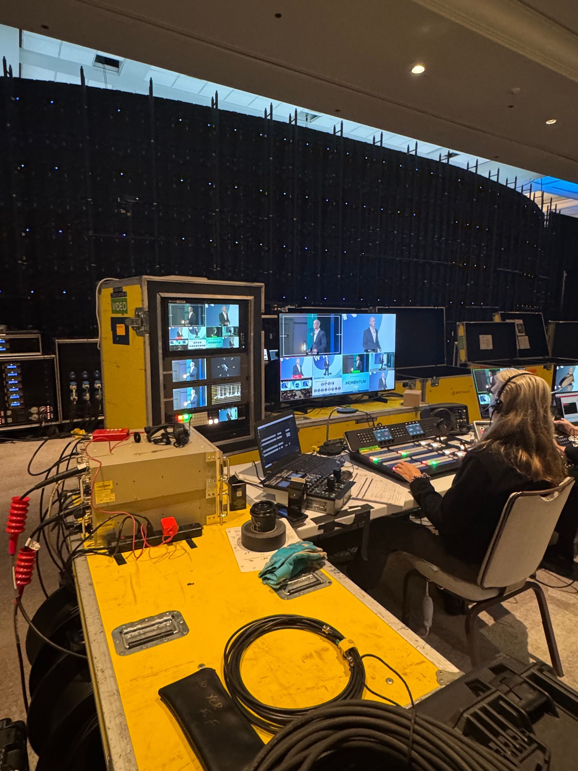 A person operates a professional video control station with multiple monitors and a mixing console in a dark hall.