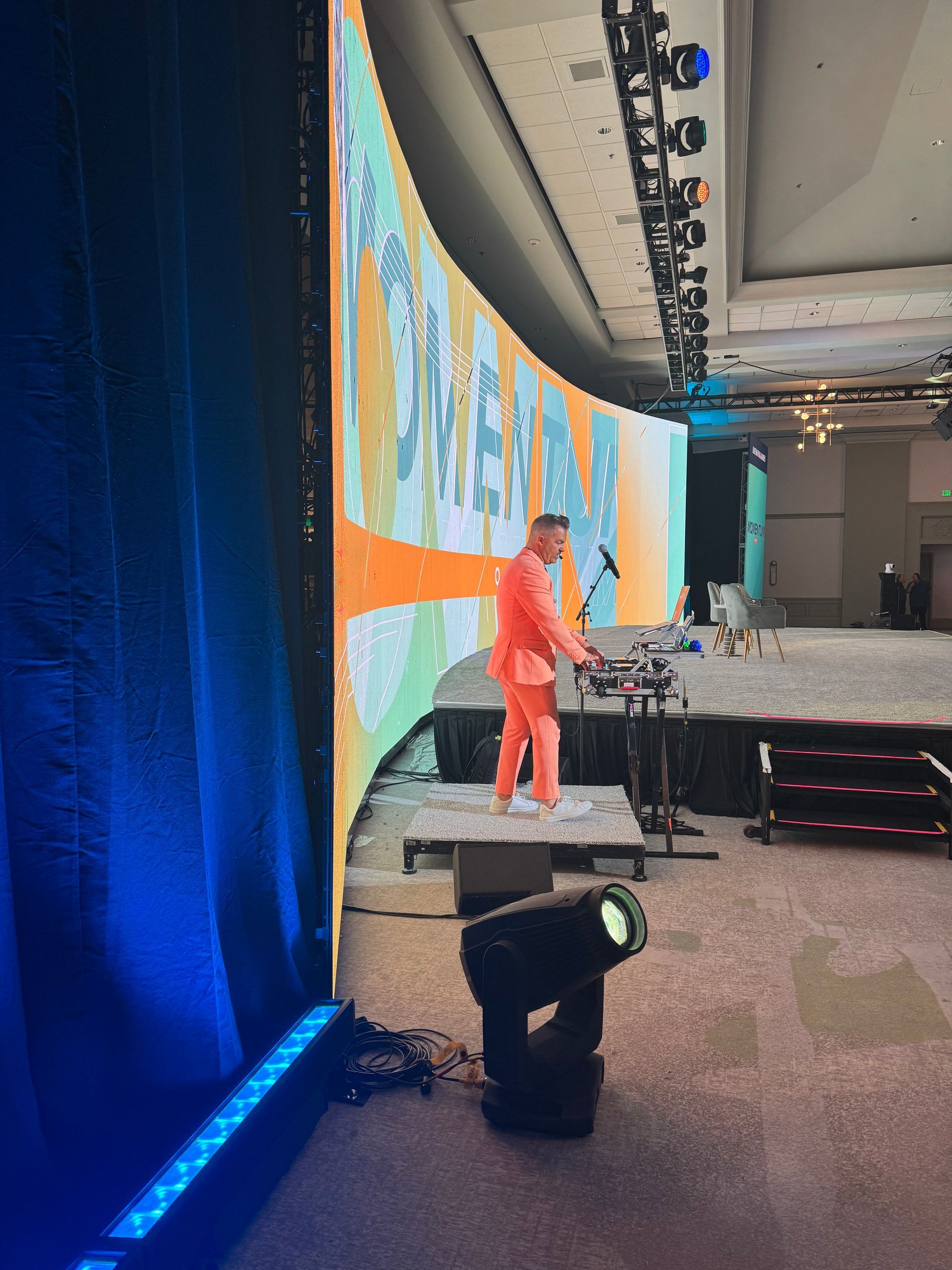 A person in a bright orange suit stands on a stage performing music in front of a large, vibrant, abstract graphic display.