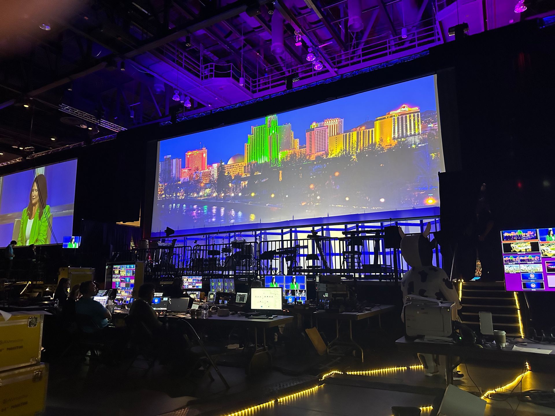A control room looking toward a large projection screen displaying a colorful cityscape, with a presenter on smaller screen.
