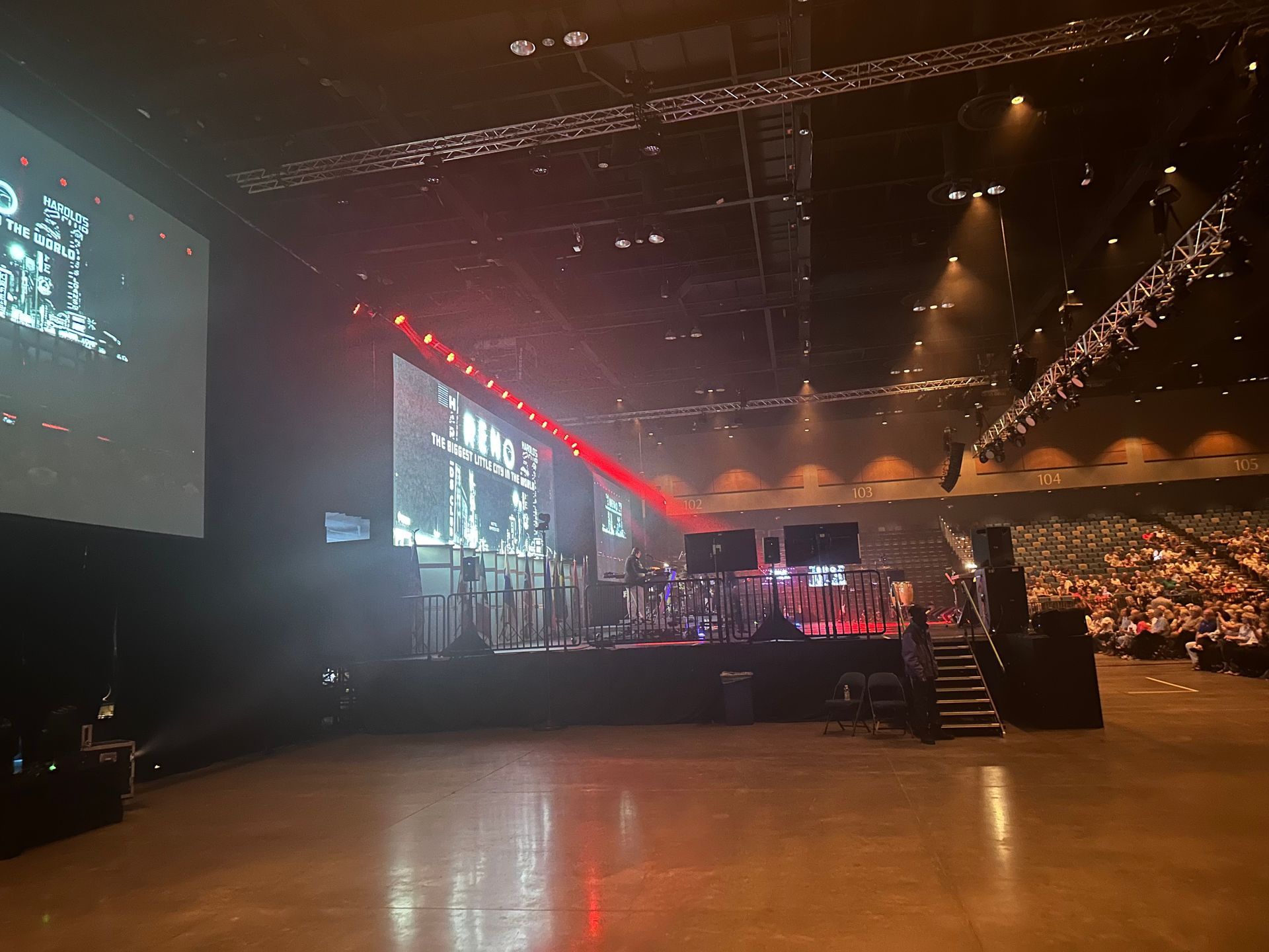 A large, dimly lit indoor event venue with a stage, screens, and auditorium seating.