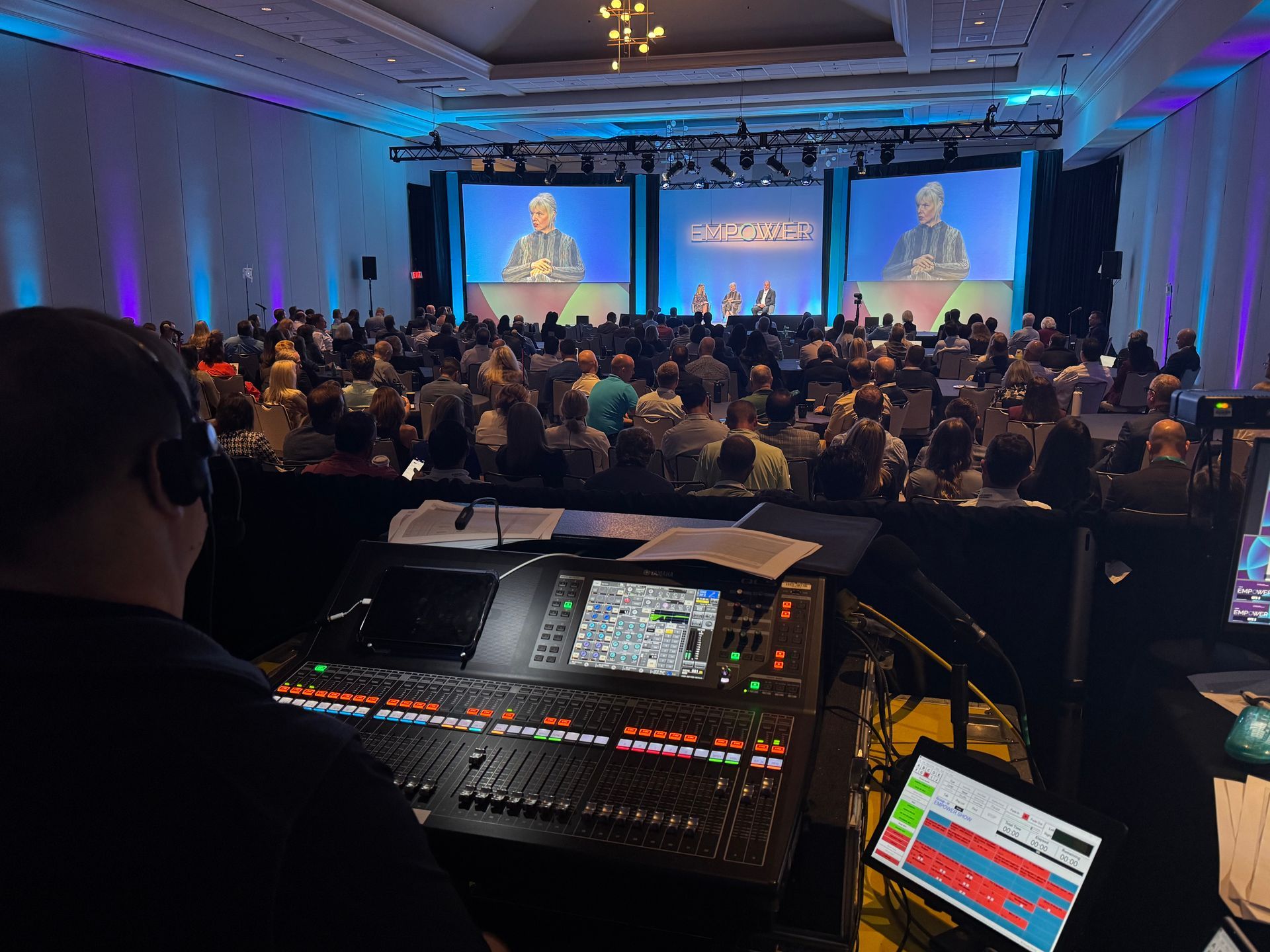 A technician operates an audio mixing console in a darkened conference hall with a large, occupied audience and stage.