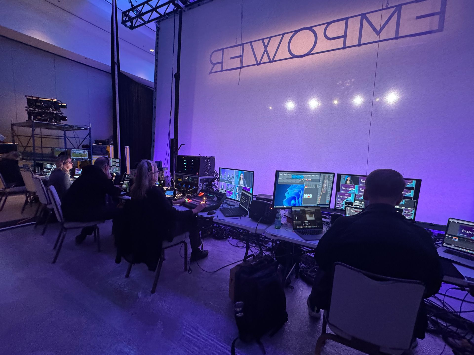 A production crew monitors screens at a technical control station in a room with a large, lit 