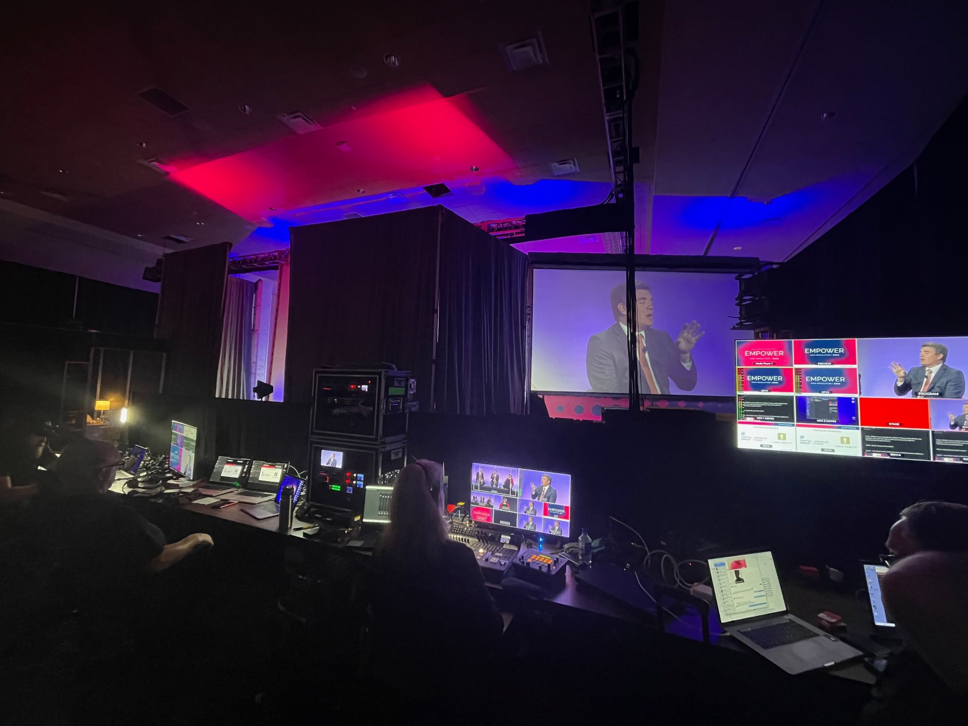 A darkened production control room with monitors displaying live broadcast feeds of a person speaking on stage.