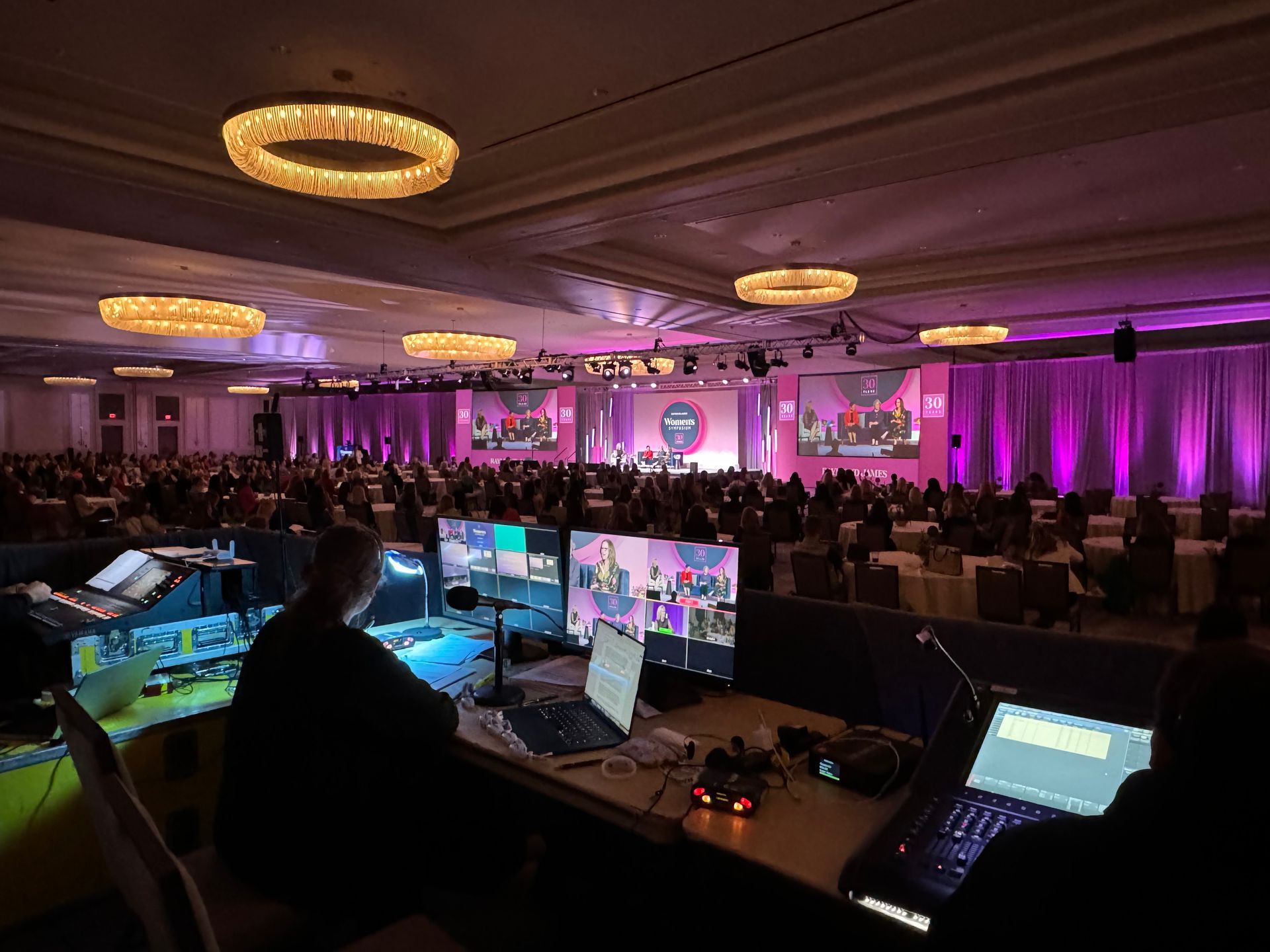A technical crew at a console manages a large, dimly lit event hall filled with attendees and purple-lit stage screens.