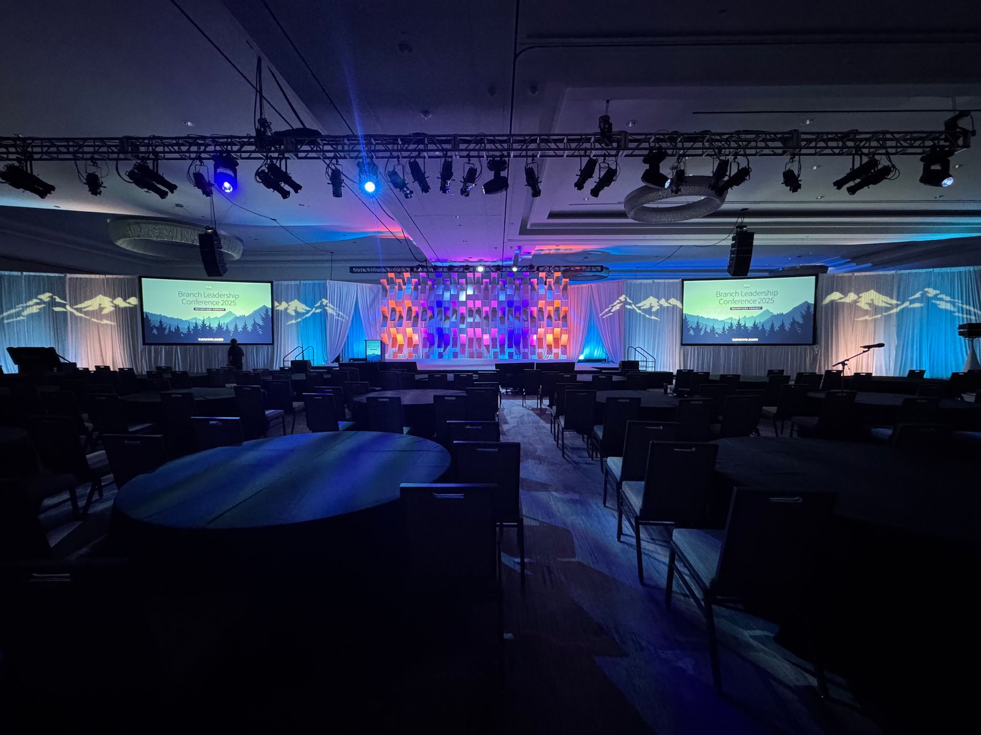 An event ballroom with round tables facing a stage illuminated with blue and purple lights and two large screens.