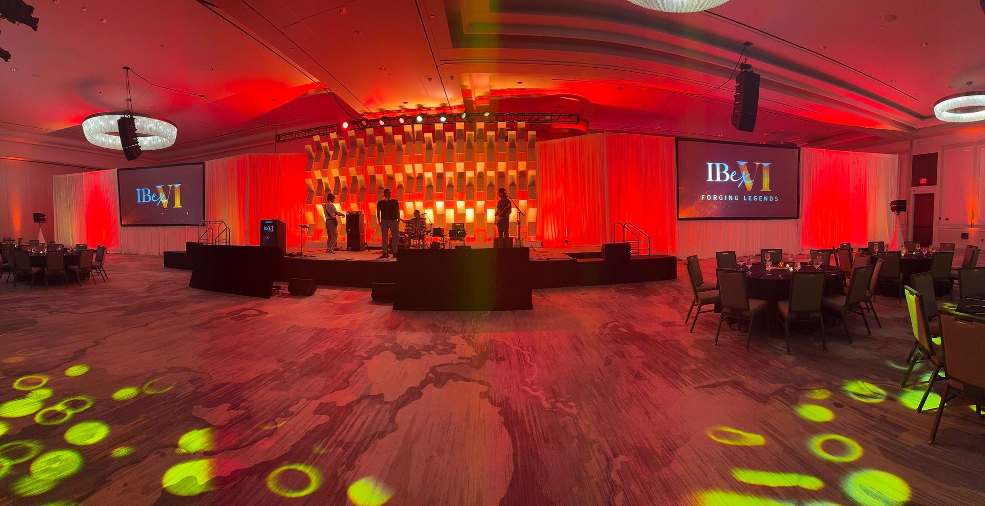 A large, dimly lit ballroom set for an event with red ambient lighting, a center stage, and circular tables for guests.