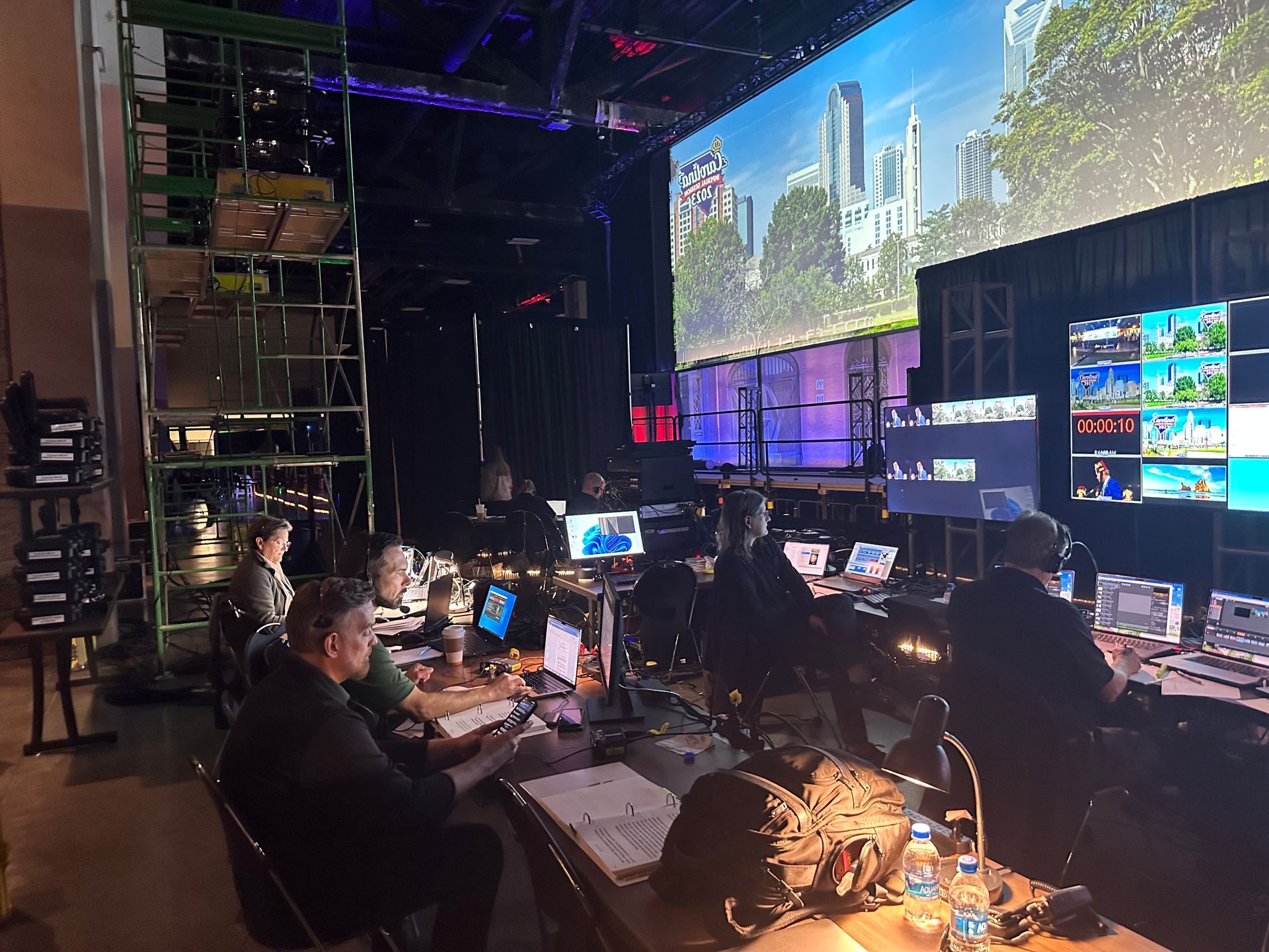 A technical production team monitors multiple screens and equipment in a darkened studio with a large city backdrop.