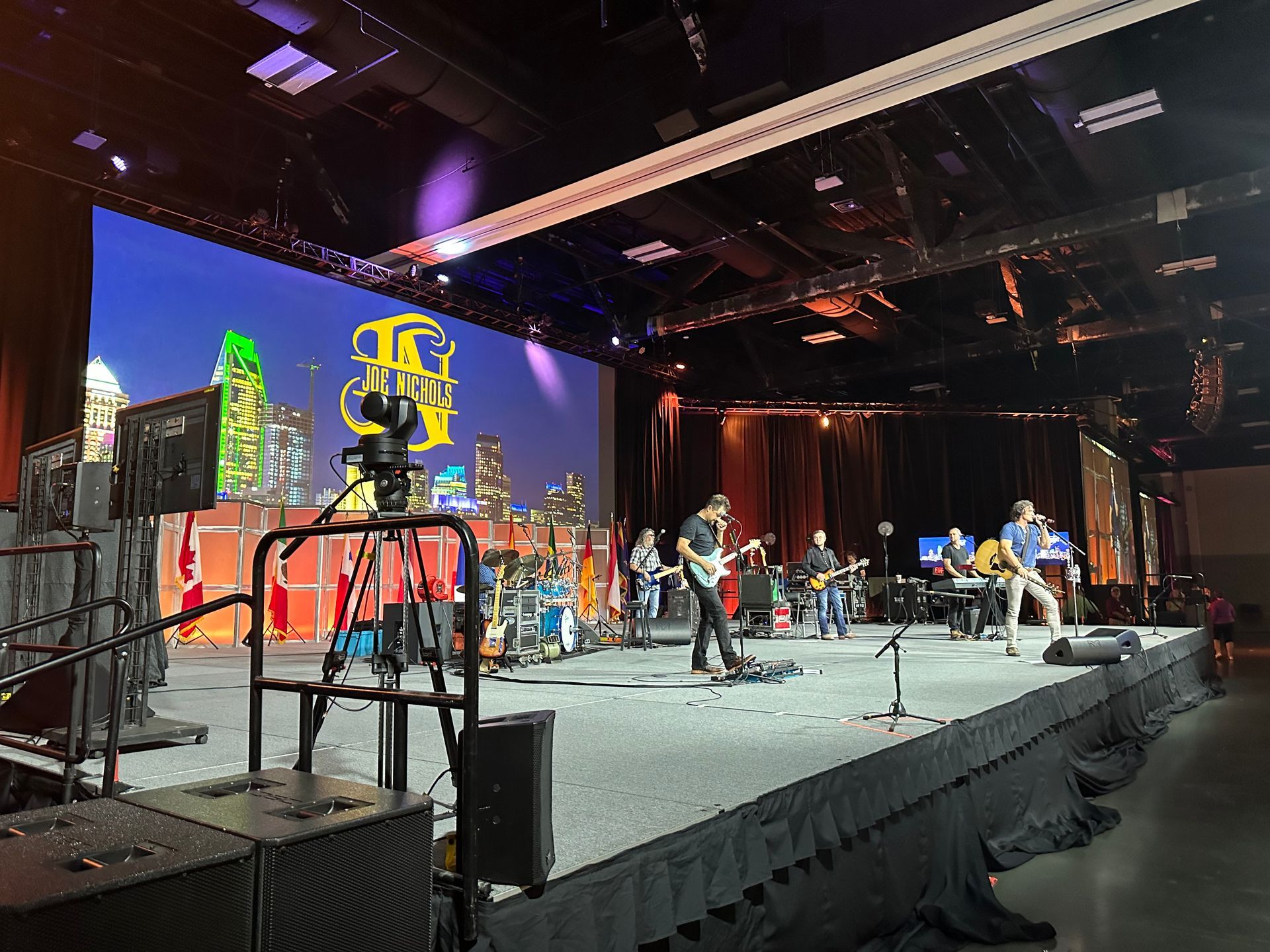 A band performs on a stage in front of a large screen displaying a stylized cityscape at an indoor event.