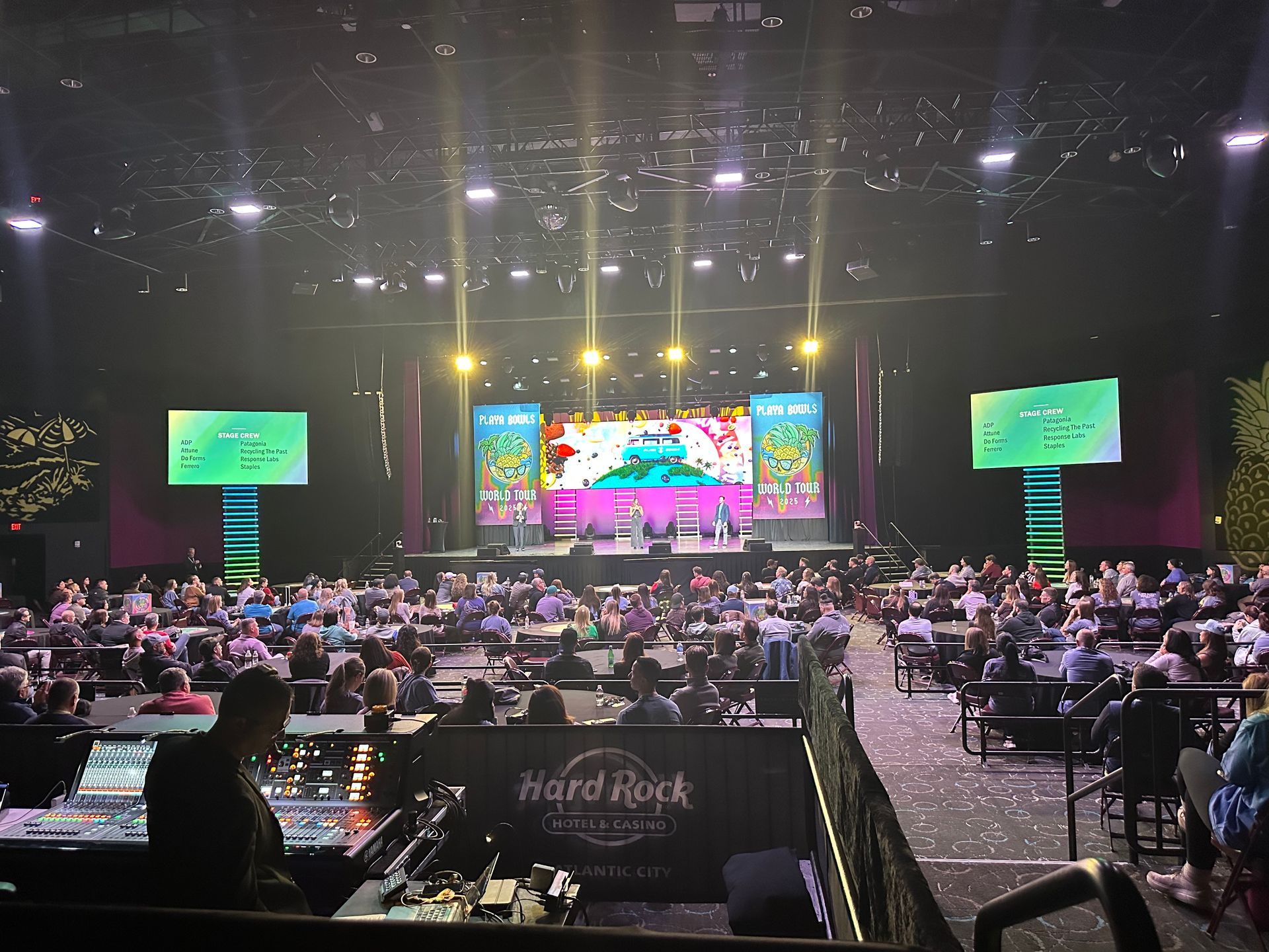 Audience members sit at tables in a dimly lit Hard Rock event hall facing a large stage with bright screens and lights.