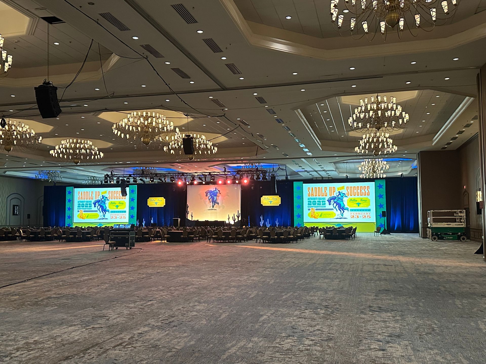 A large, empty hotel ballroom featuring a stage with three bright screens and elegant chandeliers hanging overhead.