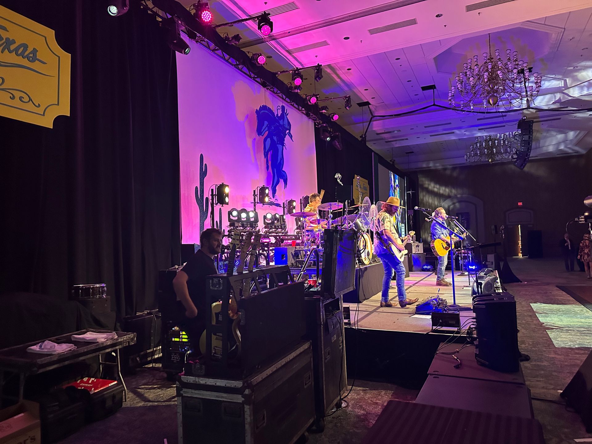 A band performs on a stage with purple lighting in a large event room, featuring a large screen and musical equipment.