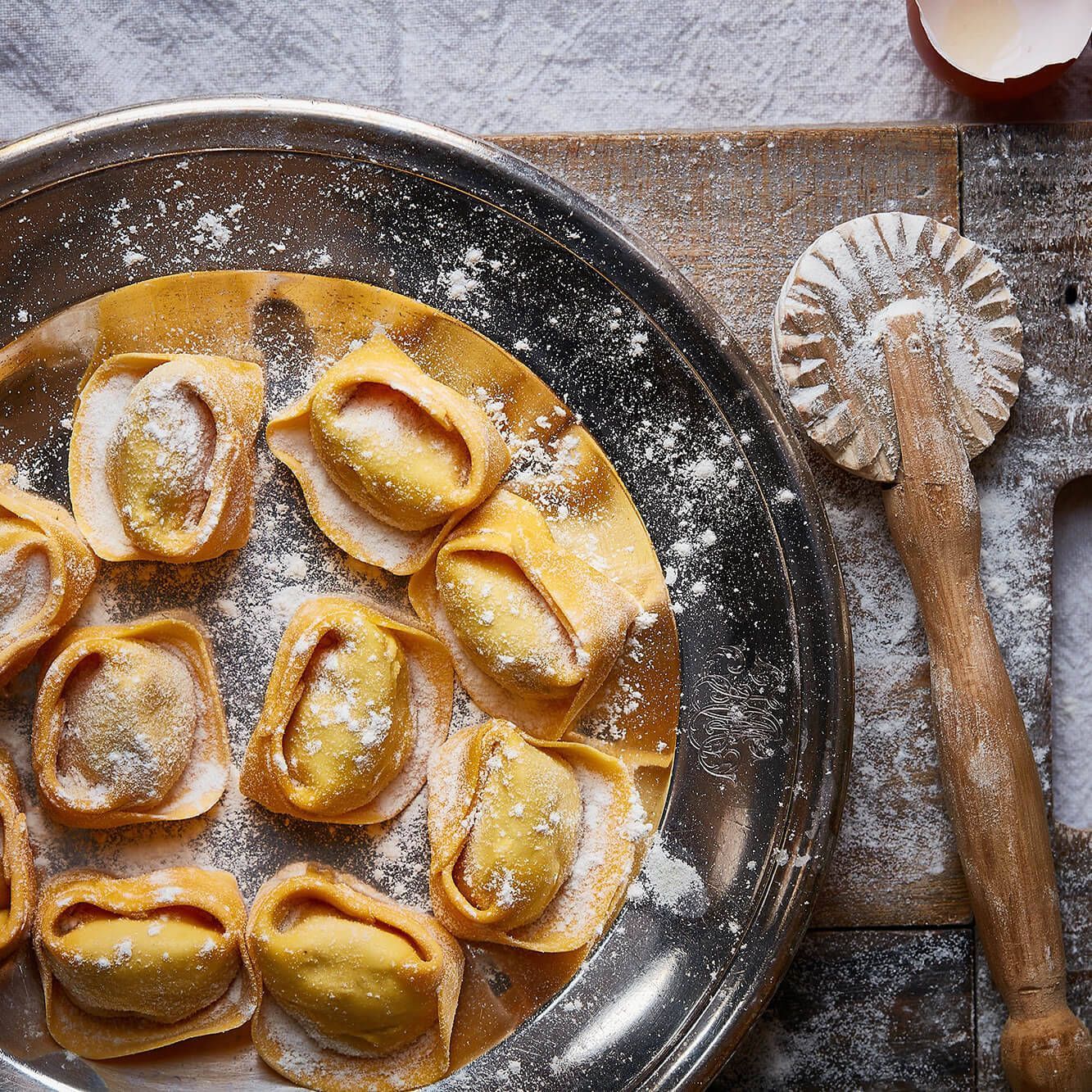 Homemade ravioli on a dark plate, dusted with flour, beside a wooden pasta wheel on a rustic surface