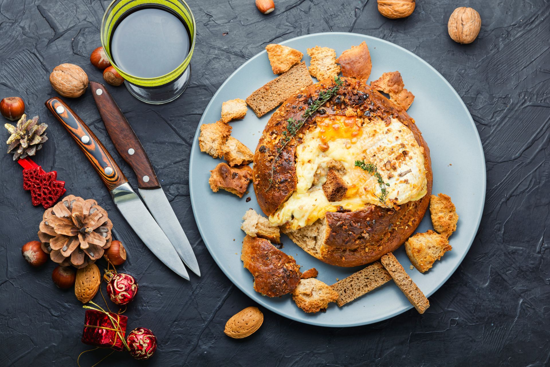 Rustic bread bowl with creamy soup, croutons, knife, and festive pinecones on a dark table.