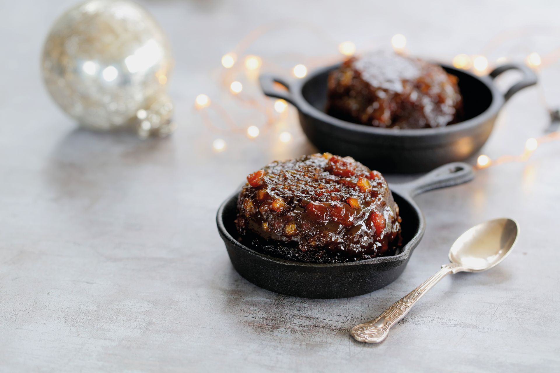Two glazed chocolate desserts in black dishes with a spoon on a light tabletop, softly lit with fairy lights