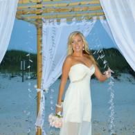 Woman in a white dress standing under a beach canopy holding flowers, with curtains and ocean in the background
