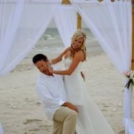 Couple under white draped canopy on a beach, posing for a romantic photo.