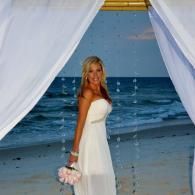 Woman in a white dress holding flowers, standing under draped fabric by the ocean at sunset