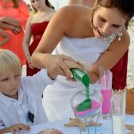 Woman in a white dress pours green liquid into a pink glass at a decorated table.