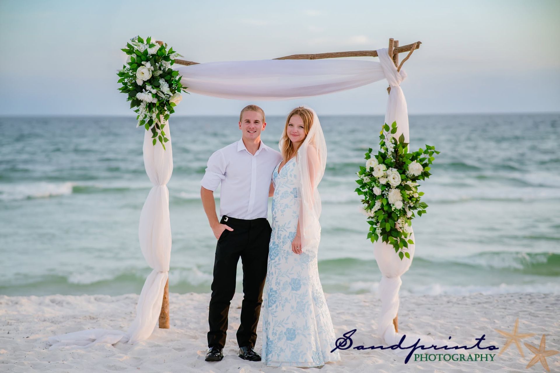 Couple standing under a floral arch on a beach with ocean waves in the background