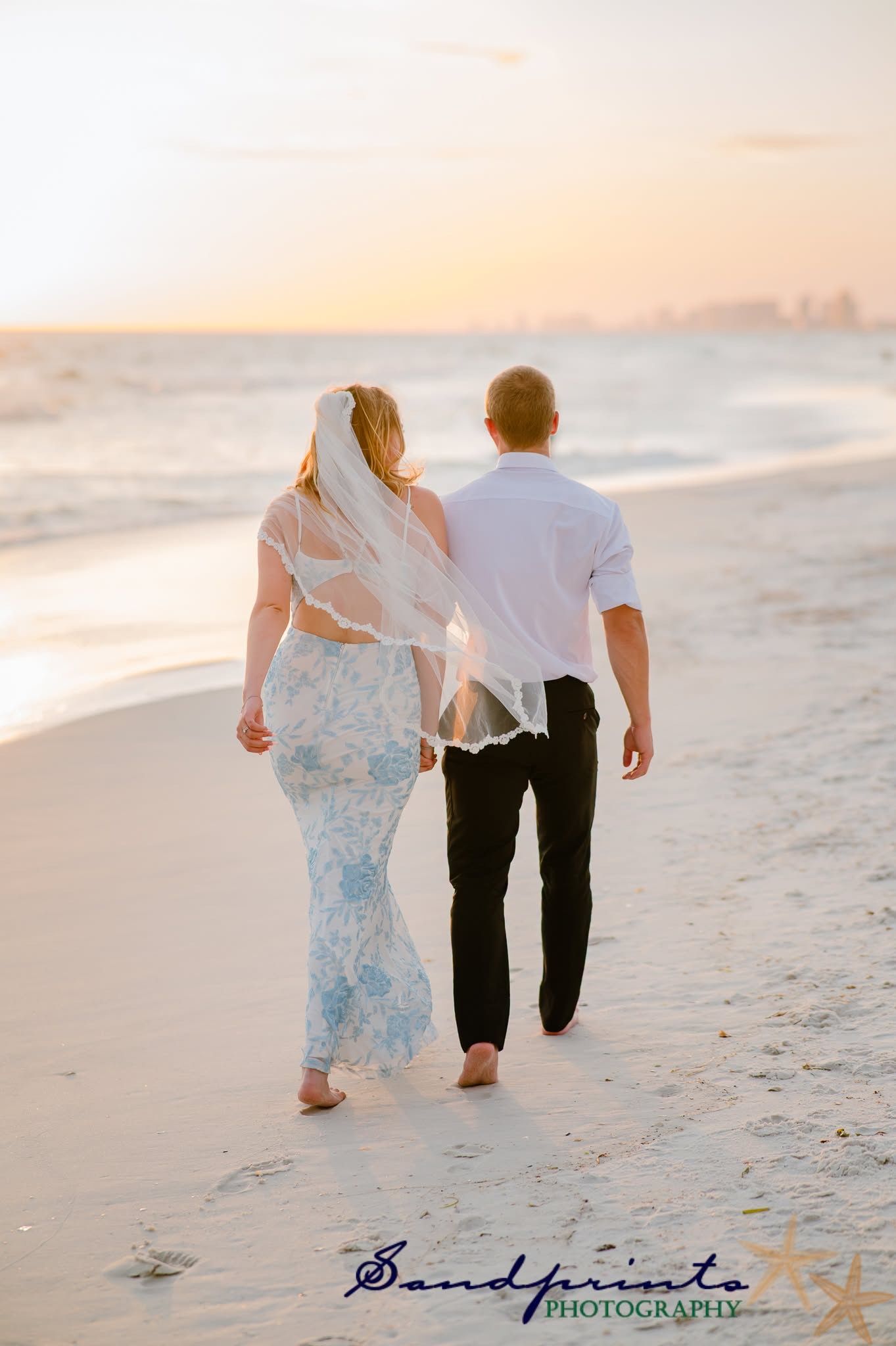 Couple walking barefoot on a beach at sunset, with a white dress and veil flowing in the breeze.