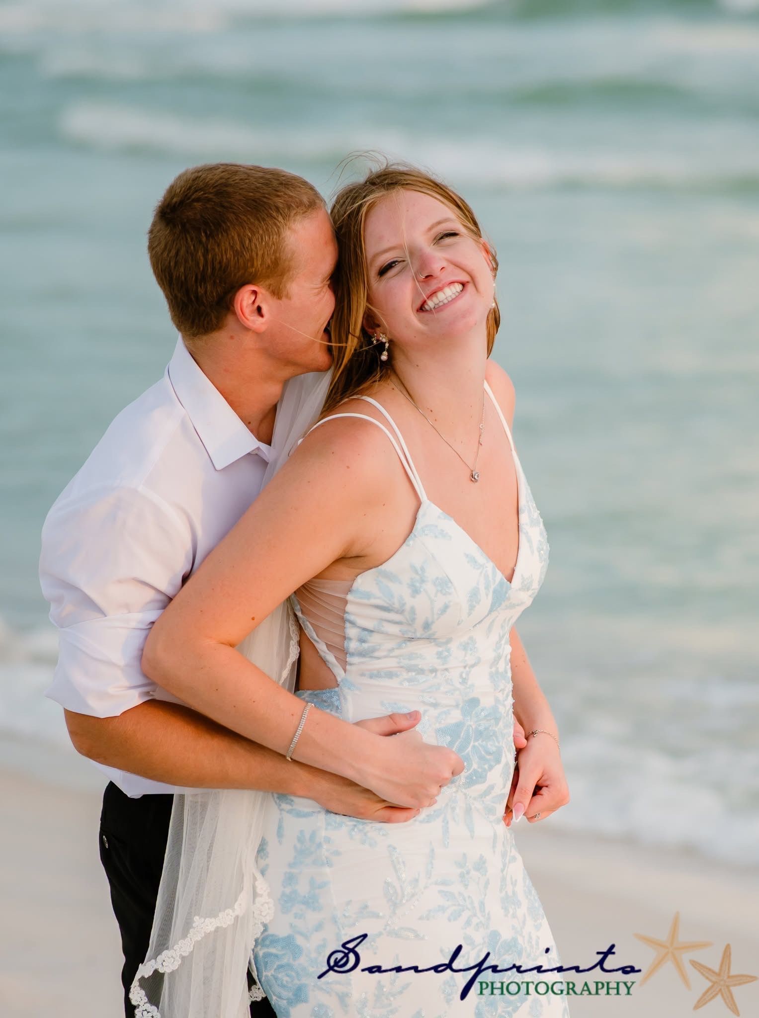 Couple embracing on a beach at sunset, smiling with waves in the background.