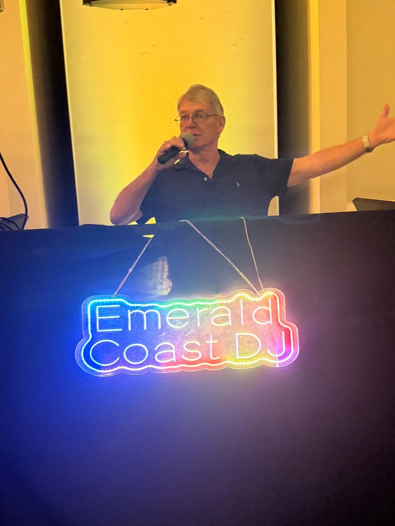 DJ speaking behind a glowing “Emerald Coast DJ” booth with one arm raised in a dim event space
