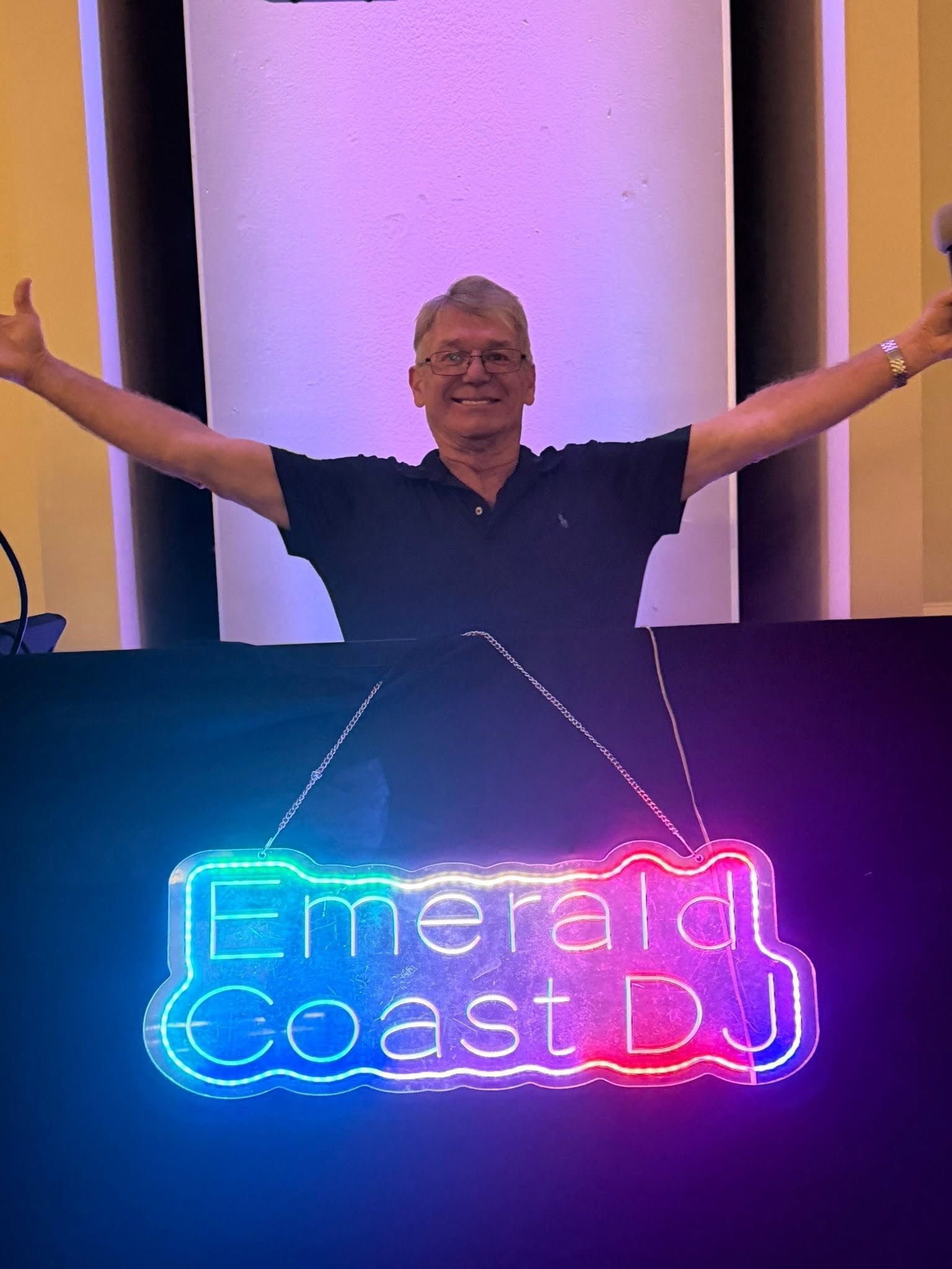 Smiling man with arms outstretched behind a neon “Emerald Coast DJ” sign at an event booth