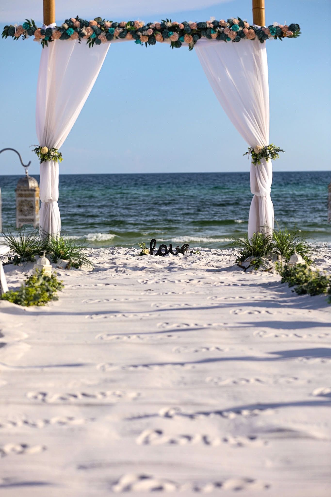 Beach wedding arch with white drapes and flowers on a sandy path facing the ocean