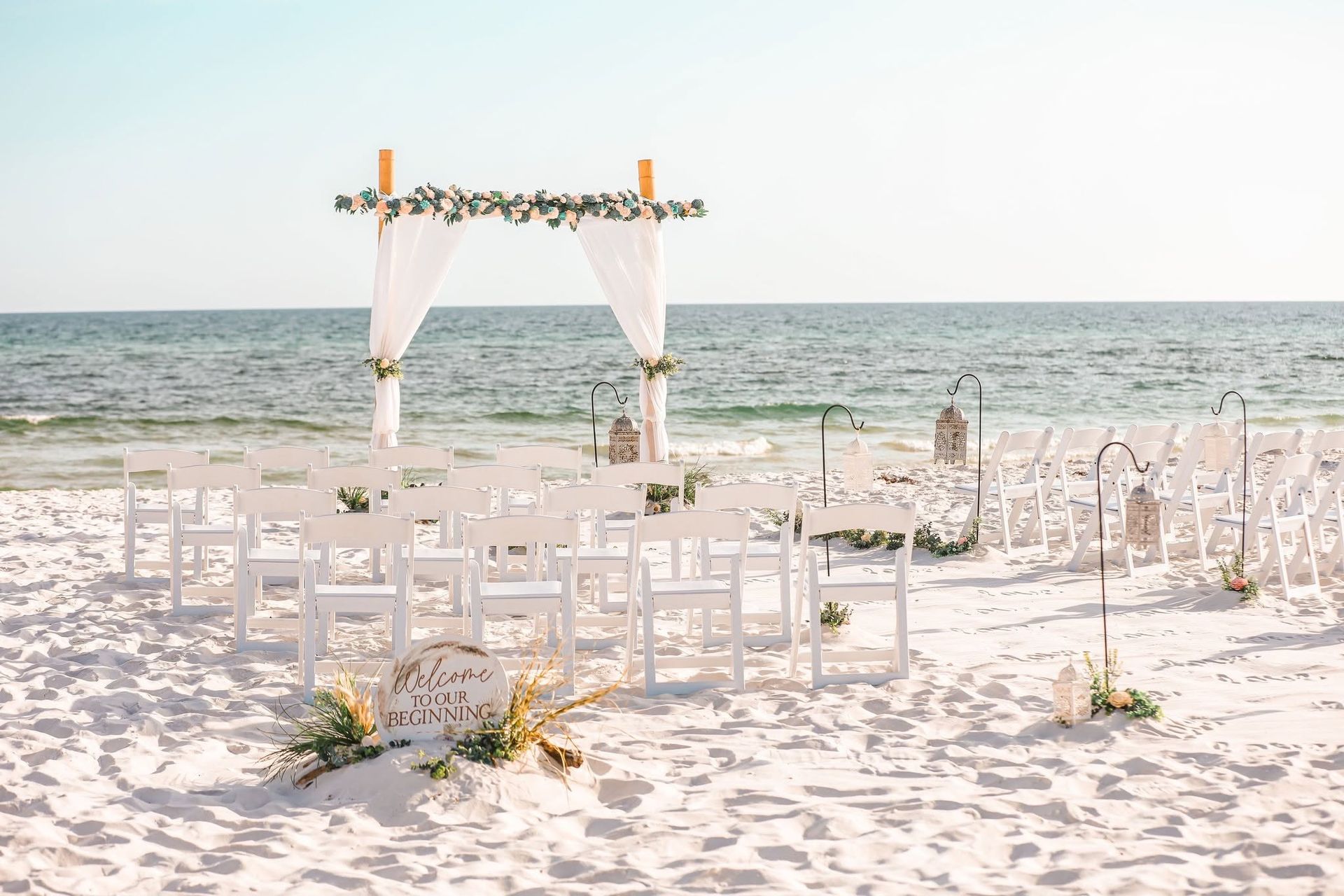 Beach wedding setup with white chairs and a floral arch beside the ocean