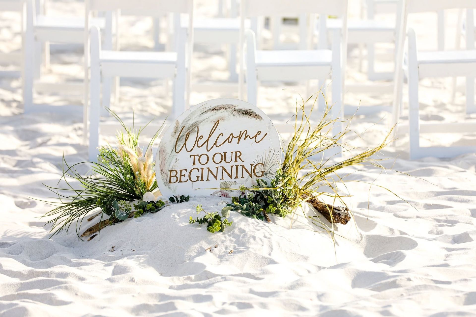 Sand sign reading “welcome to our beginning” among white chairs on a sandy beach