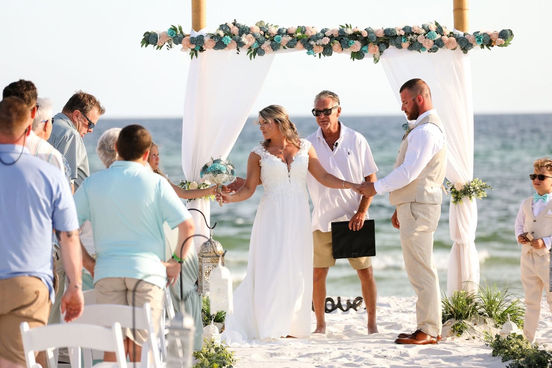 Beach wedding ceremony under floral arch with couple holding hands as guests watch nearby